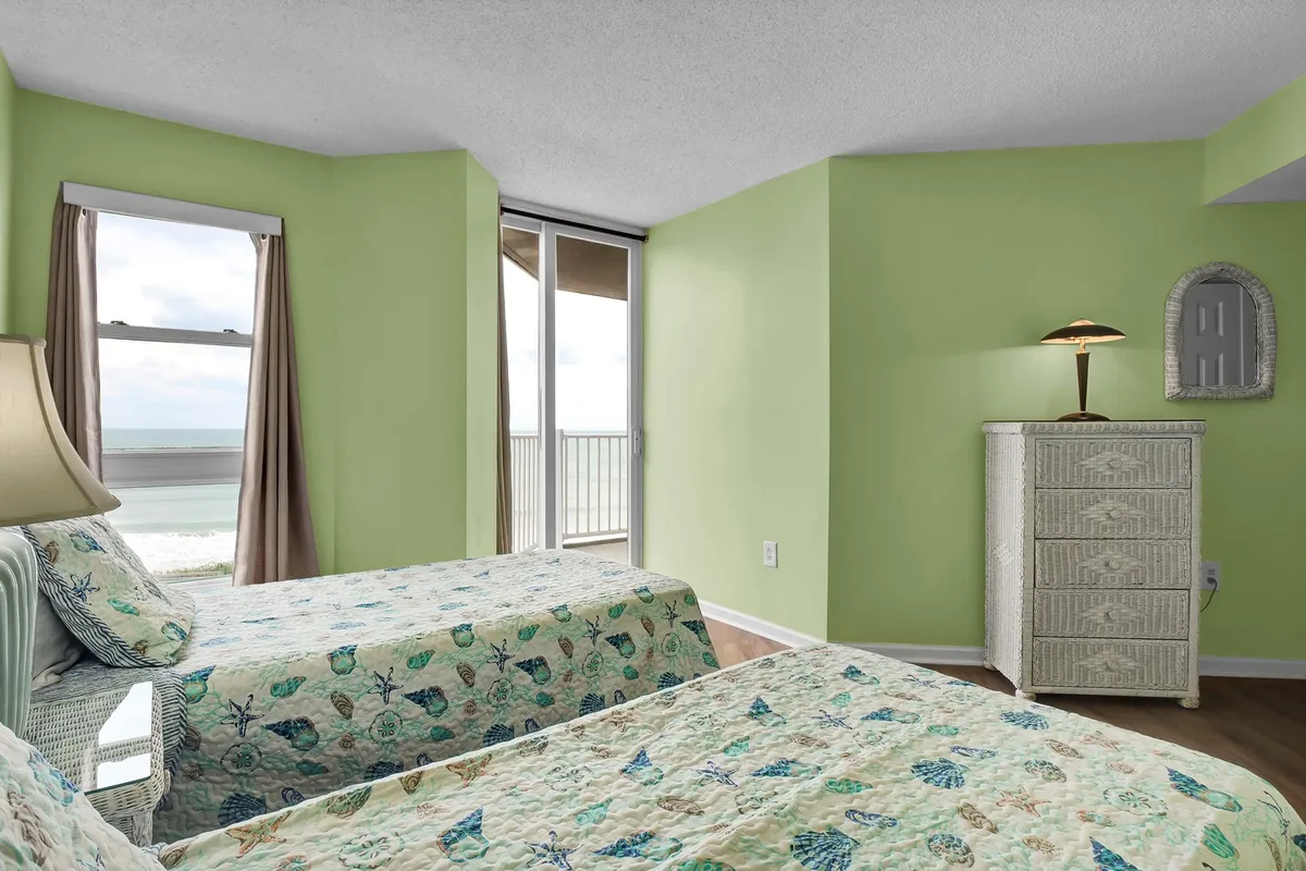 A cheerful twin bedroom with two twin beds featuring floral bedding, a large window with ocean views, a decorative lamp, and a woven dresser. The space exudes a shabby chic vibe, perfect for kids or friends seeking their own spot.