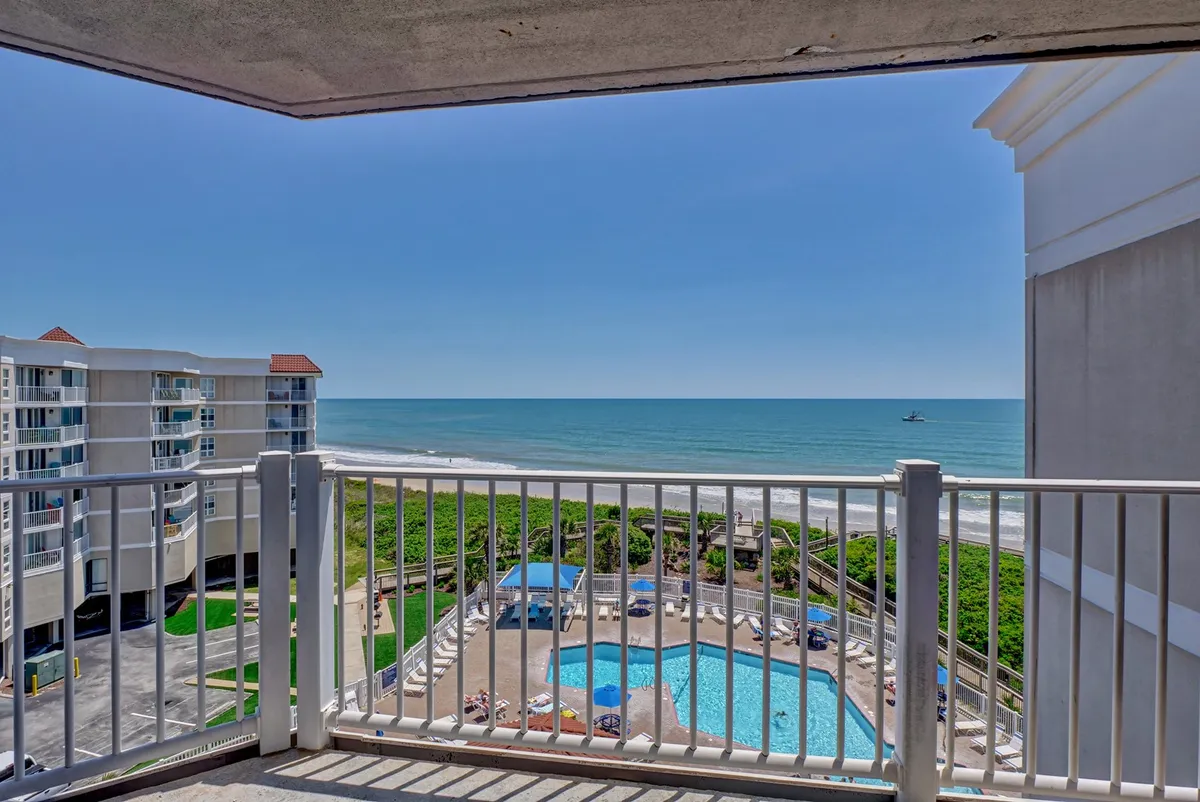 The view from this image includes a tranquil swimming pool with blue water, an ocean vista beyond, and a balcony railing in the foreground. The scene evokes leisure, with options for swimming, sunbathing, or simply taking in the view from the balcony.