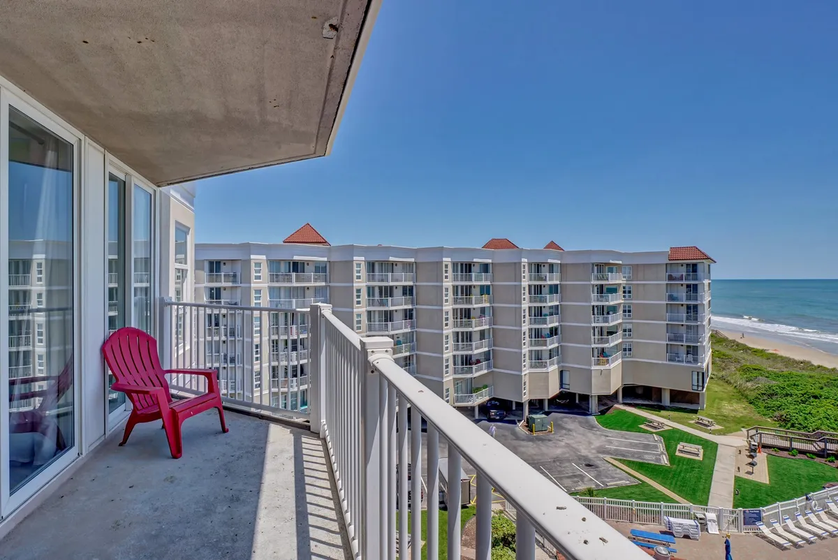This image shows a balcony with a red chair overlooking a sweeping ocean view, with the soft hues of a rising or setting sun visible on the horizon. The balcony has a sturdy railing and is surrounded by other buildings, highlighting the communal resort setting.