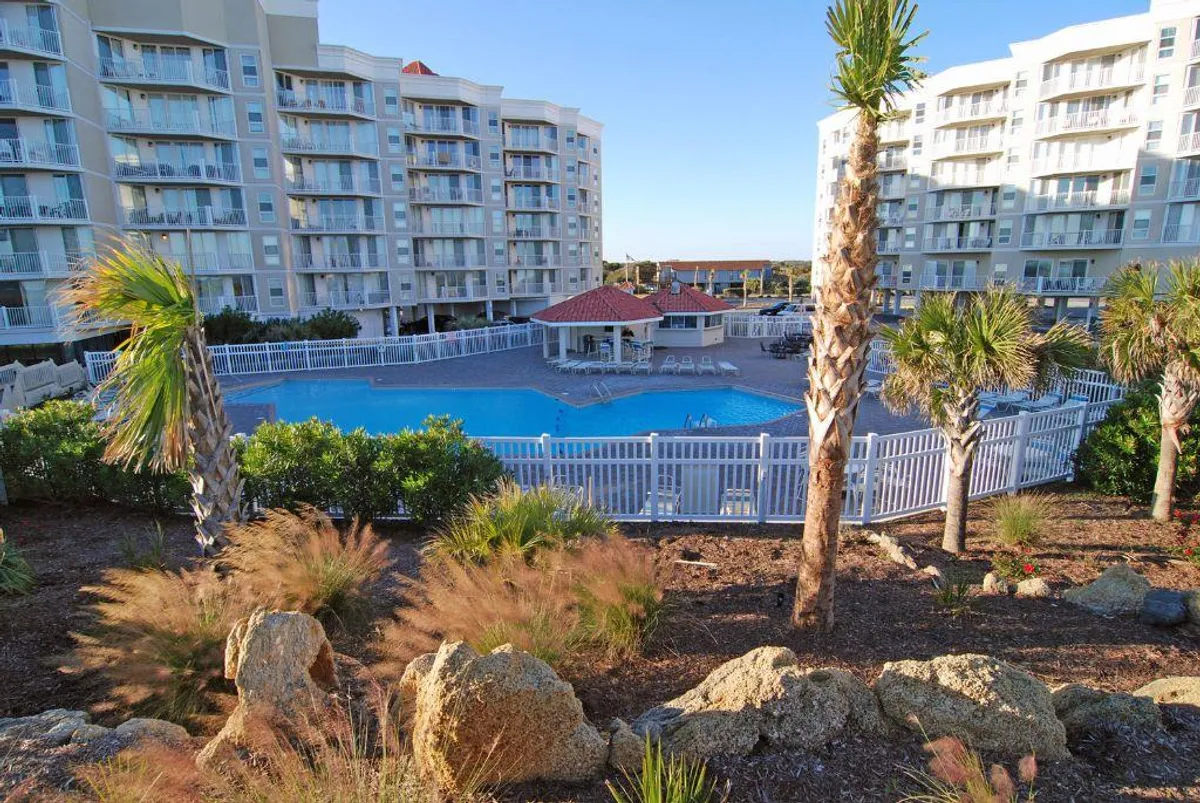 A vibrant view of the resort's swimming pool surrounded by palm trees and apartment buildings. The pool area is lively and inviting, perfect for swimming, sunbathing, and socializing with friends and family. The scene captures the essence of year-round recreation and relaxation offered by the resort.