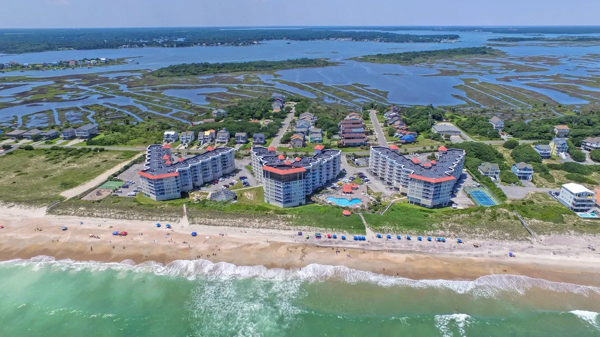 An inviting exterior scene with beachfront properties, a sparkling swimming pool, rows of colorful beach umbrellas, and gentle ocean waves in the background. The image captures families and groups enjoying swimming, sunbathing, beach volleyball, and gathering along the shoreline.
