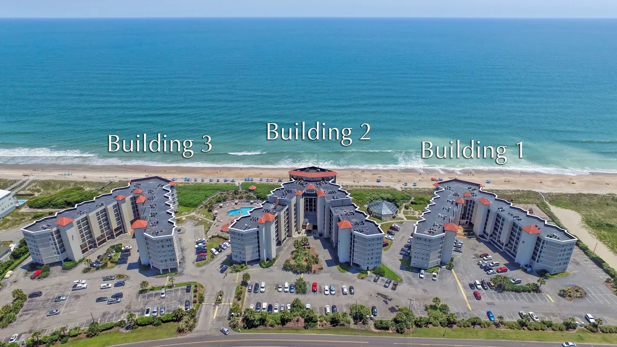 A wide-angle exterior view showing three buildings, a sparkling swimming pool, and the ocean beyond. The pool is surrounded by loungers and is positioned to maximize ocean views, with sunset light hinting at tranquil evenings. Balconies overlook the water, reinforcing the connection between accommodations and outdoor amenities.
