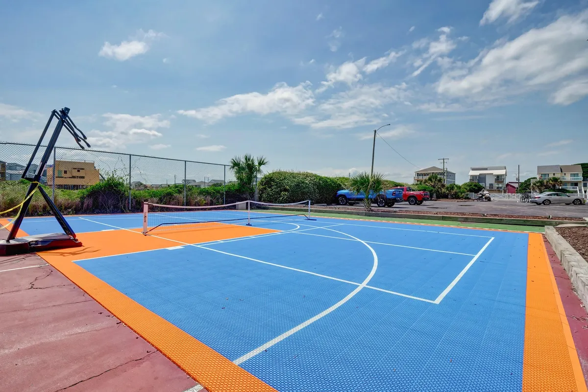 A lively outdoor recreation area featuring a basketball hoop and a tennis net on brightly colored court surfaces. The space is open and inviting, ideal for friendly sports matches and family play, with a warm, playful mood.