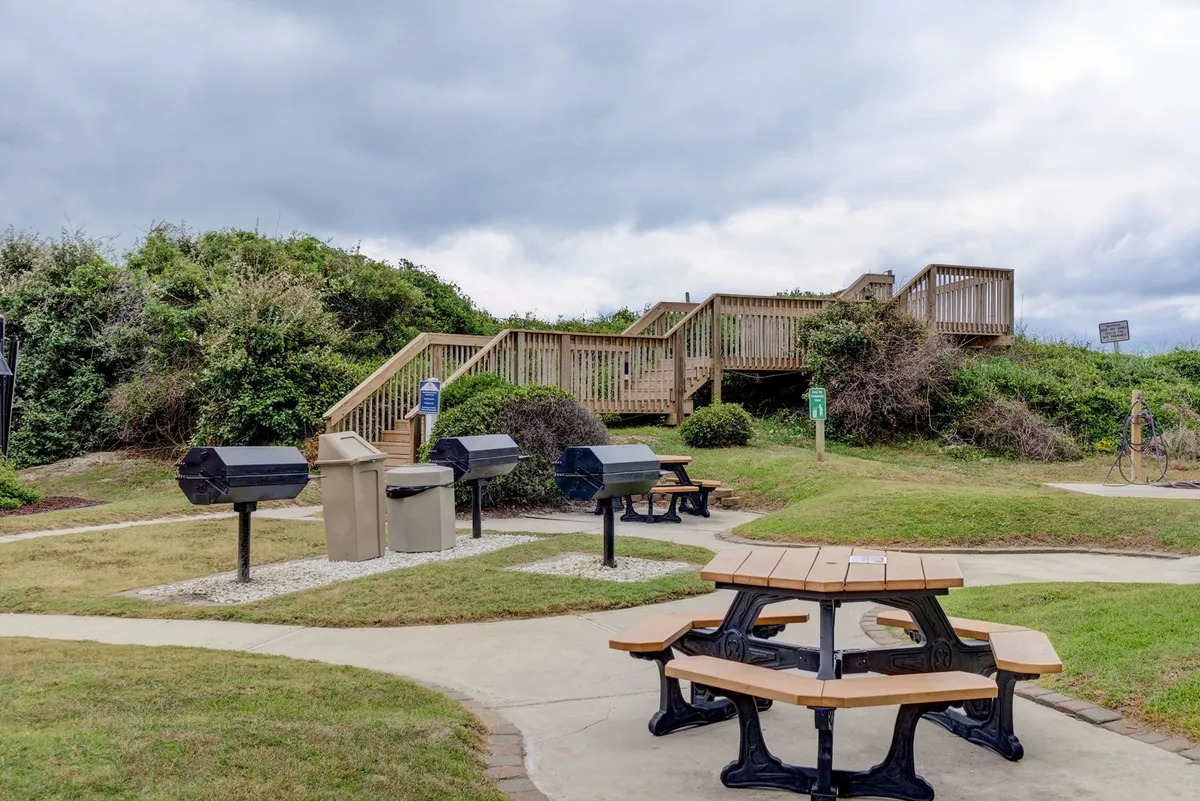 An inviting outdoor grilling and picnic area featuring picnic tables, built-in grills, and a staircase leading to a raised vantage point. The scene is set for group gatherings or intimate meals, with the option to watch the sunset and enjoy fresh, self-catered dinners outdoors.