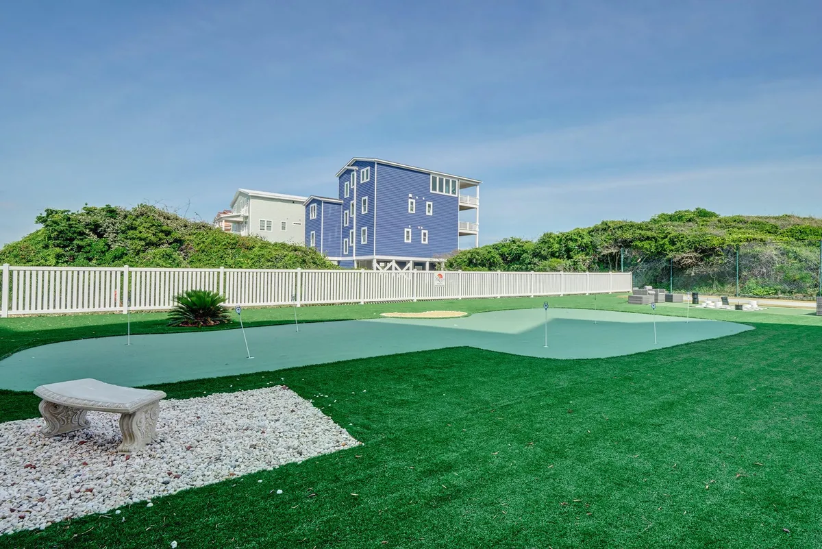 This exterior image features a vibrant green artificial turf area, a blue house in the background, and a decorative stone bench. The open outdoor space suggests opportunities for relaxation, light recreation, or informal gatherings with sunset views over the lawn.
