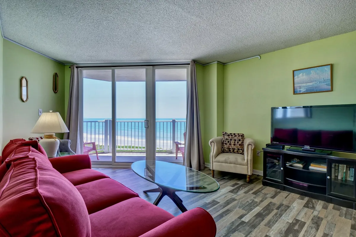 A cozy living room with a red sofa, glass coffee table, TV unit, and wide sliding doors that frame a sweeping ocean view. Natural light pours in, illuminating the comfortable seating and creating a warm, inviting atmosphere perfect for relaxing or socializing.