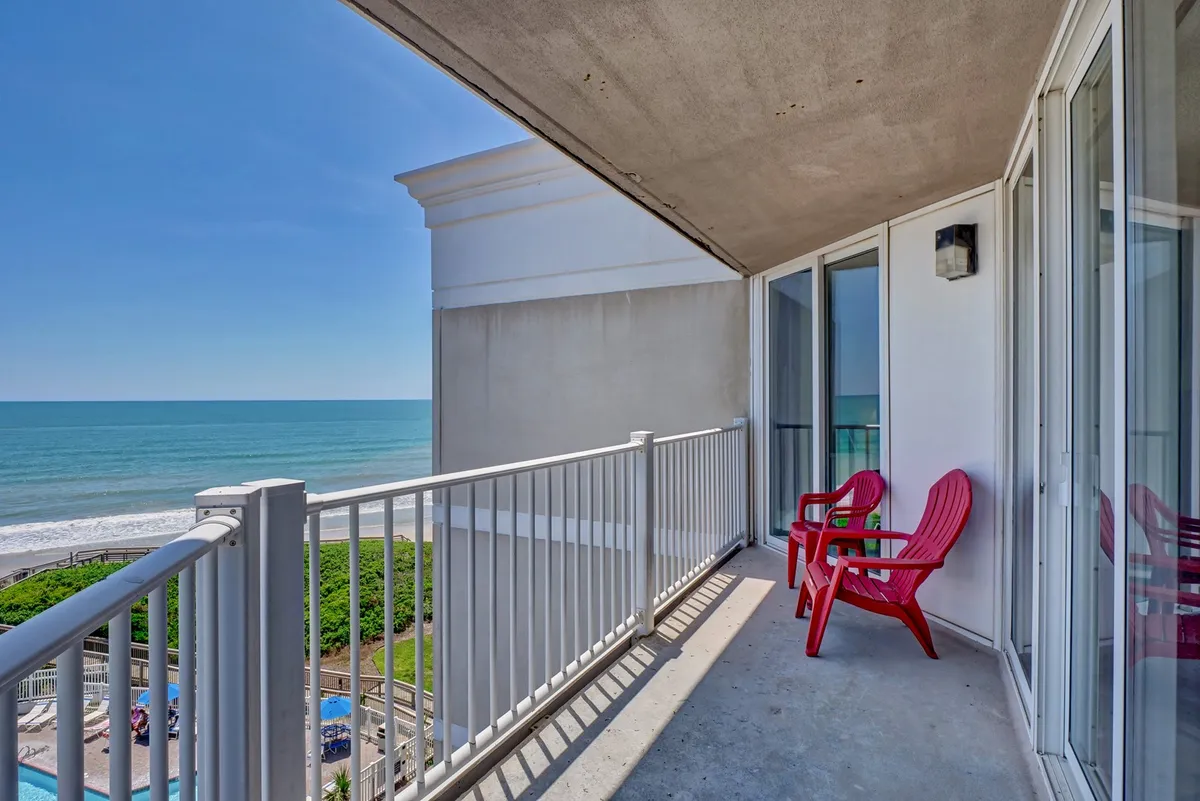 A serene oceanfront balcony with red Adirondack chairs facing the sea, bordered by a glass railing. The balcony invites relaxation, sunrise coffee, and deep breaths of fresh ocean air, embodying the indoor-outdoor lifestyle described in the section.